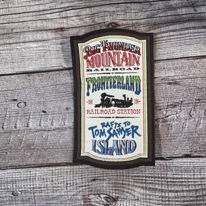 Może przedstawiać: Naszywka w stylu vintage z kolorowym wzorem z tekstem "Big Thunder Mountain Railroad Frontierland Railroad Station Rafts to Tom Sawyer Island".