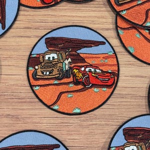 Könnte beinhalten: Ein runder gestickter Aufnäher mit Lightning McQueen und Mater aus dem Disney Pixar Cars Film. Der Aufnäher zeigt die beiden Charaktere, die durch eine Wüstenlandschaft mit einer großen Felsformation im Hintergrund fahren. Der Aufnäher ist perfekt, um Ihren Besitztümern einen Hauch von Cars-Filmmagie zu verleihen.