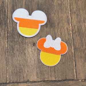 Pode incluir: Dois patches de feltro para passar a ferro em forma de cabeça do Mickey Mouse. Um é laranja e amarelo com contornos brancos, e o outro é laranja com um laço branco e contornos brancos.
