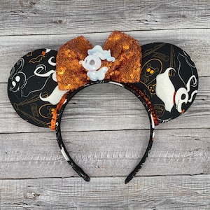 May include: Black and white mouse ears with a black and white patterned fabric featuring the text "Zero" and a white ghost with a black outline. The ears have an orange sequined bow with a white ghost charm.