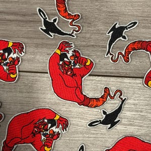 May include: Embroidered patches featuring a red genie character with yellow accents and a black genie lamp. The patches are in various poses and sizes, with white borders, suitable for clothing or accessories.