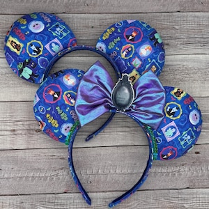 Puede incluir: Un par de orejas de Minnie Mouse azules y moradas con un lazo morado iridiscente y un amuleto de la Mansión Encantada. Las orejas están cubiertas de una tela azul con un patrón de personajes de la atracción de la Mansión Encantada.