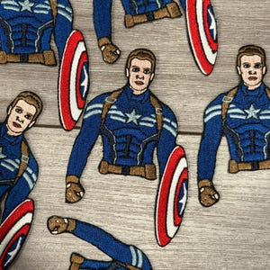 May include: Embroidered patches featuring Captain America in a blue and brown uniform with a white star on the chest. The patches include the Captain America shield, red with white stripes and a blue star.