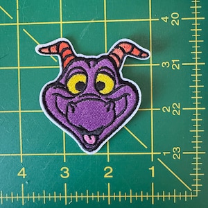 May include: A purple embroidered patch featuring a cartoon character with yellow eyes, a smiling mouth, and red horns. The patch is approximately 2 inches wide and 2 inches tall.