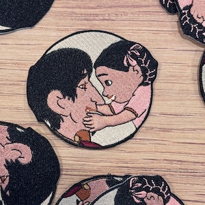 May include: Embroidered patch depicting a parent and child in a loving embrace. The design shows a parent with dark hair and a child in a pink outfit. The patch has a white border and is suitable for clothing or accessories.