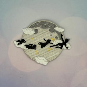 May include: Embroidered patch featuring a gray moon with a textured surface, white clouds, and black silhouettes of figures in flight. Yellow stars are scattered across the design. The patch is circular with a light beige border.