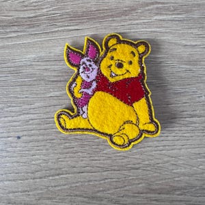 以下が含まれることがあります： くまのプーさんとピグレットが刺繍されたパッチ。プーさんは黄色で赤いシャツを着ています。ピグレットはピンクで、プーさんに抱っこされています。