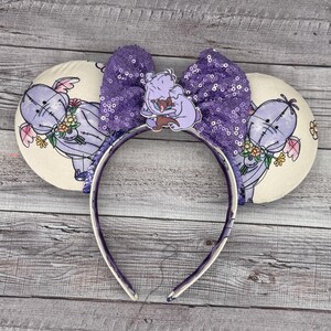 Puede incluir: Orejas de Minnie Mouse de lentejuelas moradas con un diseño de Dumbo y su madre en morado y blanco en las orejas.