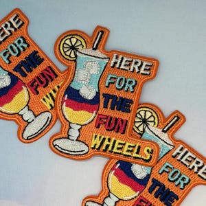 May include: Embroidered patches in orange, featuring a cocktail design with the text "HERE FOR THE FUN WHEELS". The cocktail has blue, red, and yellow layers, a lemon slice, and a straw. The patches are ideal for jackets, bags, or other fabric items.