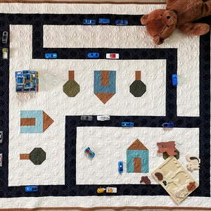 May include: A white and cream-colored quilt with a black road design, toy cars, and building blocks. The quilt features a road map design with toy cars and building blocks. A brown teddy bear is resting on the quilt. A wooden puzzle is also on the quilt.