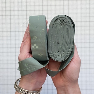 Könnte beinhalten: Zwei Rollen aus salbeigrünem Stoffband werden gezeigt. Das Band hat ein sich wiederholendes Muster aus kleinen weißen Kreuzen und Strichen. Eine Rolle ist flach, die andere eng gewickelt.