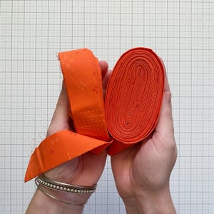 Può includere: Due mani che mostrano strisce di tessuto arancione. Una mano tiene strisce piegate con un motivo a croce, l'altra un rotolo. Il tessuto è arancione brillante e lo sfondo è una griglia. Adatto per il patchwork.