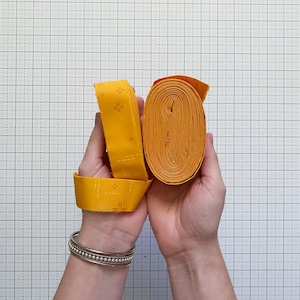 May include: Two rolls of yellow fabric strips are held in hands. One roll is tightly wound, while the other is partially unrolled, revealing a repeating pattern of small cross-like designs. The fabric is a vibrant yellow color.