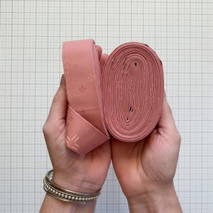 May include: Two hands holding a roll of pink fabric strips. The fabric has a repeating pattern of small, stylized cross-like designs. The fabric is rolled into a compact oval shape, with the end of the strip folded over.