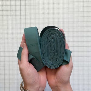 May include: A roll of teal-colored fabric ribbon with a stitched cross design. The ribbon is held in two hands, showcasing its texture and detail. The background is a grid pattern.