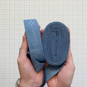 Könnte beinhalten: Zwei Hände halten eine Rolle blauen Stoffes und ein Stück desselben Stoffes. Der Stoff hat ein weißes Punktmuster und weiße Nahtlinien. Der Stoff ist hellblau.