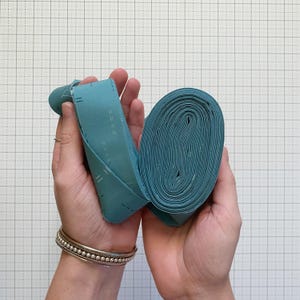 Puede incluir: Un rollo de tela color turquesa sostenido en dos manos. La tela es de color liso con un sutil patrón cosido. La tela está enrollada en espiral compacta, con una parte desenrollada, revelando el detalle cosido. El fondo es una cuadrícula.