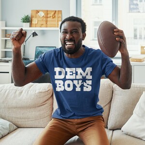 May include: A man wearing a blue t-shirt with the text "DEM BOYS" in white letters is sitting on a couch and holding a brown football. He is smiling and appears to be excited.