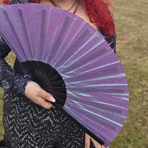 May include: A person holds a purple glitter fan with black ribs. The fan has a shimmering, iridescent finish. The person has bright red curly hair and is wearing a dark top and patterned trousers. The background is a blurred outdoor scene.