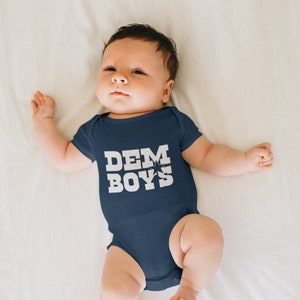 Pode incluir: Um beb&ecirc; vestindo um macac&atilde;o azul marinho com o texto "DEM BOYS" em letras brancas.