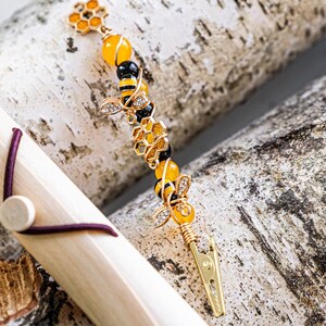 K&ouml;nnte beinhalten: Eine Holzkiste mit einem braunen Gummiband und einem goldenen Bienen-Anh&auml;nger mit gelben und schwarzen Perlen. Der Anh&auml;nger ist an einem goldenen Clip befestigt.