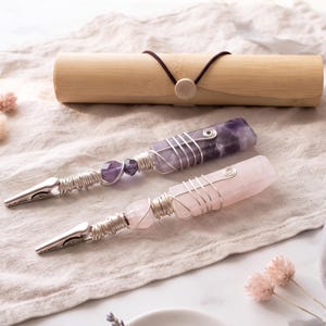 May include: Two metal roach clips featuring raw crystal handles wrapped in silver-toned wire. One clip has a deep purple amethyst stone, while the other features a pale pink rose quartz stone. A cylindrical bamboo storage case is included.