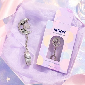 May include: A silver cigarette holder with a crescent moon and star design, and a dangling ring. The holder is presented in a lavender box, which reads "MOON CIGARETTE HOLDER". The box also includes the text "silver moon + dangling star" and "sparkle up your ritual".