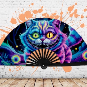Op de afbeelding: Decoratieve handventilator met een levendige illustratie van de Cheshire Cat uit Alice in Wonderland. De ventilator toont de iconische grijns van de kat, met een kleurenpalet van blauw, paars en geel. De ventilator heeft een zwarte handgreep.