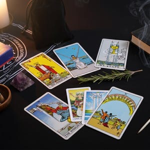 Puede incluir: Un conjunto de cartas del tarot con ilustraciones coloridas, incluyendo El Mago y El Colgado, se disponen sobre una superficie negra. También hay una caja, un libro, una vela y una pluma, sugiriendo un tema místico o espiritual.