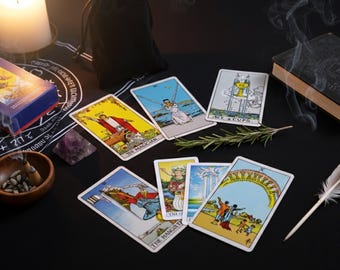 May include: A collection of tarot cards with colorful illustrations, including The Magician, The Hanged Man, and Ace of Cups, are arranged on a black surface. A box, a book, a candle, and a feather are also present, suggesting a mystical or spiritual theme.