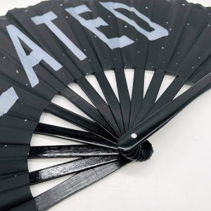 以下が含まれることがあります： 白い「LATER」のデザインが施された黒い折りたたみ式の扇子。扇子は閉じられており、持ち手がみえています。