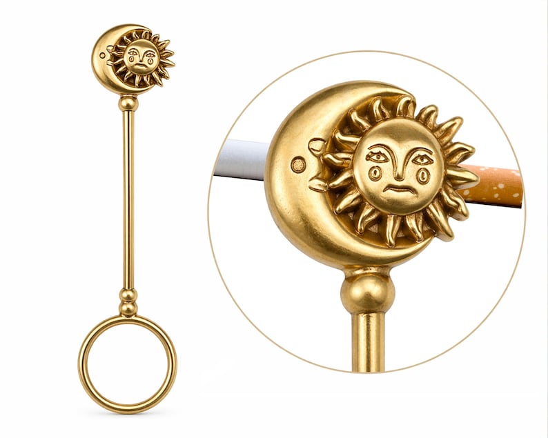 May include: A gold-colored cigarette holder featuring a sun and moon design. The holder has a ring for the finger and a long stem. The sun and moon are detailed with facial features. The holder is designed to hold a cigarette.