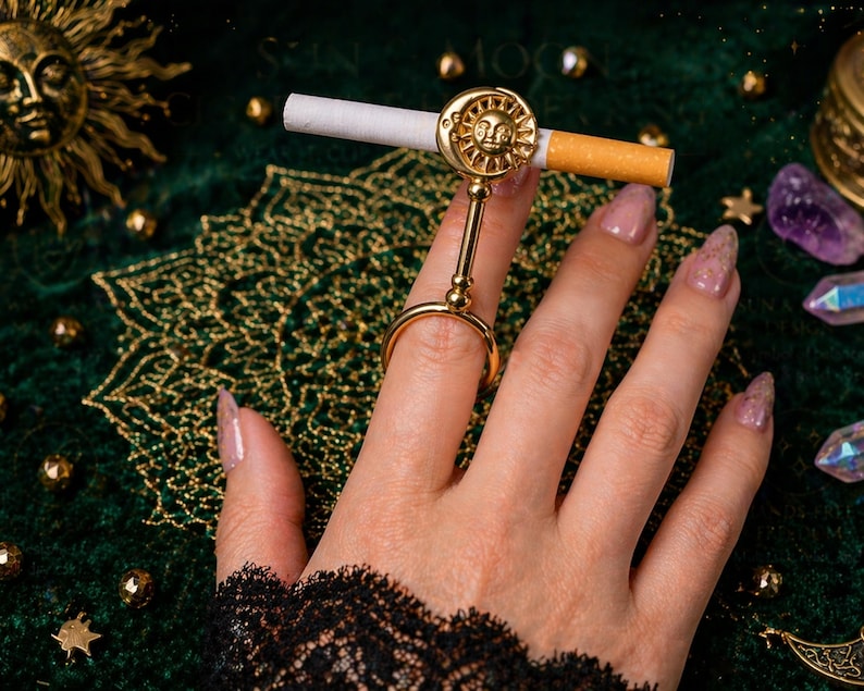 May include: A gold-colored cigarette holder ring with a sun design, holding a lit cigarette. The ring is worn on a finger, set against a dark green background with gold accents. The sun design is detailed and ornate.
