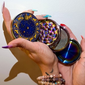 Pode incluir: Um moedor de metal de quatro peças com um design zodiacal na peça superior. O moedor tem um acabamento arco-íris iridescente.
