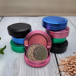 Op de afbeelding: Een set van zes ronde metalen containers met deksels. De containers zijn in verschillende kleuren, waaronder zwart, blauw, groen, roze en zilver. De deksels zijn allemaal gesloten. Eén container heeft een ontwerp met een halve maan en sterren op het deksel.