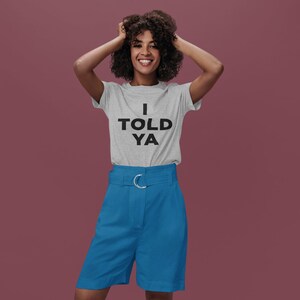 Puede incluir: Una mujer con una camiseta gris con el texto "I TOLD YA" en letras negras. Tambi&eacute;n lleva pantalones cortos azules con una hebilla de cintur&oacute;n plateada.