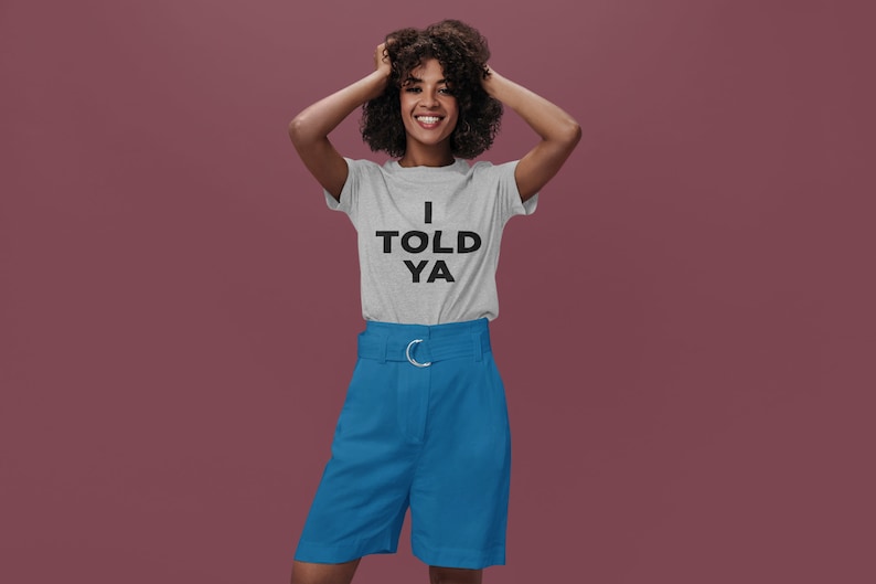 Puede incluir: Una mujer con una camiseta gris con el texto "I TOLD YA" en letras negras. Tambi&eacute;n lleva pantalones cortos azules con una hebilla de cintur&oacute;n plateada.
