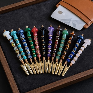 May include: A collection of twelve roach clips featuring mushroom-shaped stone toppers in various colors including white, turquoise, blue, green, red, black, purple, brown, and pink, with beaded stems and gold-toned metal alligator clips.