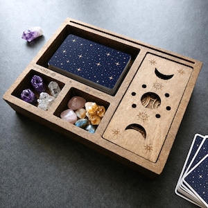 Peut inclure: Une boîte de rangement rectangulaire en bois avec plusieurs compartiments contenant un jeu de cartes bleu foncé à motifs d'étoiles, divers cristaux bruts et un couvercle coulissant gravé de phases lunaires et de symboles solaires.