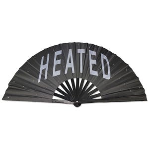 以下が含まれることがあります： 銀色のグリッターと「HEATED」という文字が印刷された黒い折りたたみ式扇子です。