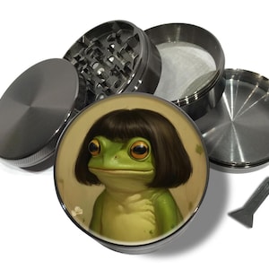 May include: A disassembled, four-piece, metal herb grinder. The top piece has a lid, and the bottom piece has a frog illustration. The frog has a green body, large eyes, and a dark brown bob haircut. A small, black scraper is included.