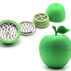 May include: A green apple-shaped herb grinder with a leaf and stem detail. The grinder is disassembled, showing the internal components, including a metal grinding surface and a mesh screen. The parts are green and silver.