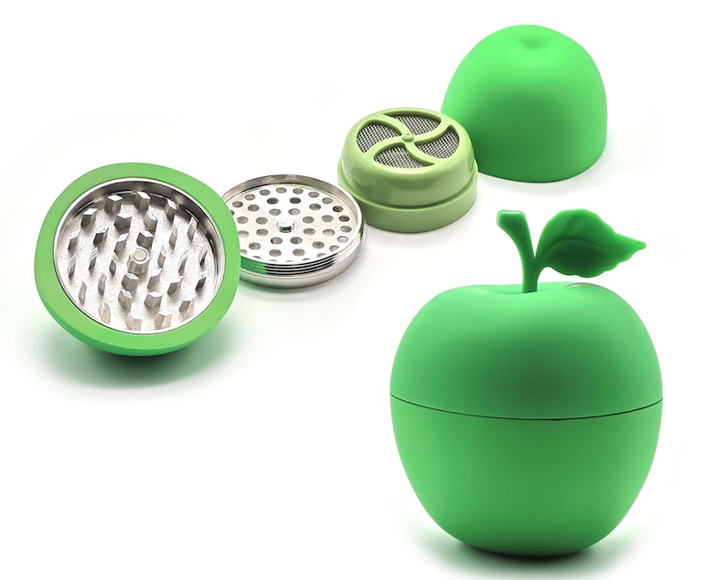 May include: A green apple-shaped herb grinder with a leaf and stem detail. The grinder is disassembled, showing the internal components, including a metal grinding surface and a mesh screen. The parts are green and silver.