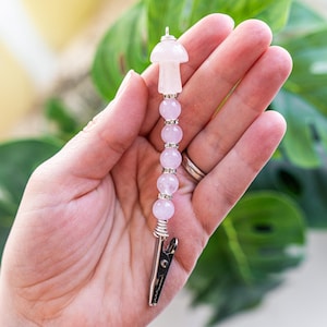 May include: A pink rose quartz mushroom charm with silver beads and a silver clip. The charm is held in a hand.