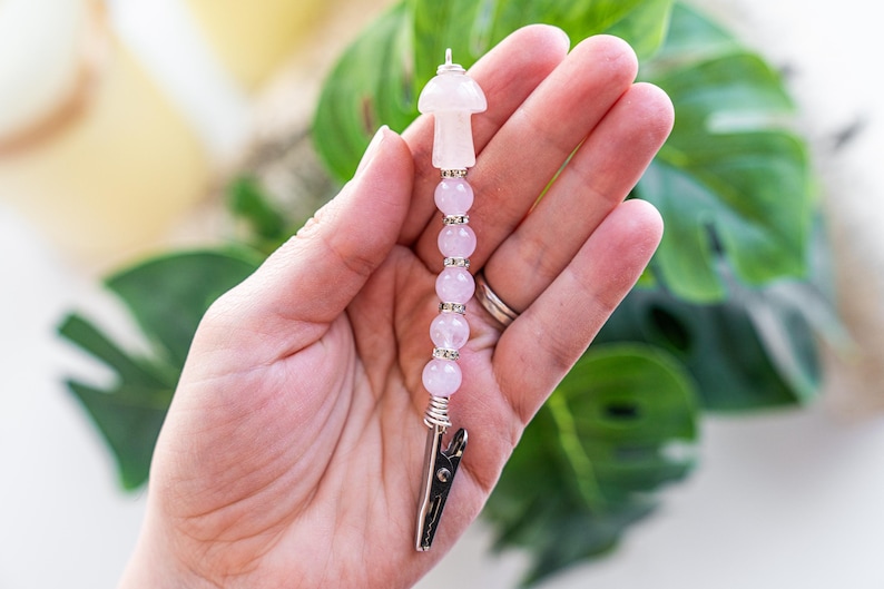 May include: A pink rose quartz mushroom charm with silver beads and a silver clip. The charm is held in a hand.