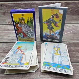 Może przedstawiać: Talia kart tarota z niebieskim pudełkiem z ilustracją królowej. Talia zawiera karty z ilustracjami "Głupca", "Królowej Mieczy" i "Asa Kielichów". Karty leżą na drewnianej powierzchni.