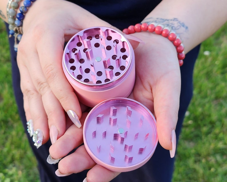May include: Two pink metal grinders with a circular design. The grinders have a series of holes in the top and bottom sections.