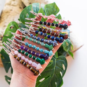 May include: A hand holding a set of 10 colorful beaded clips with silver metal clips. Each clip has a different combination of beads, including pink, blue, green, brown, and black beads.