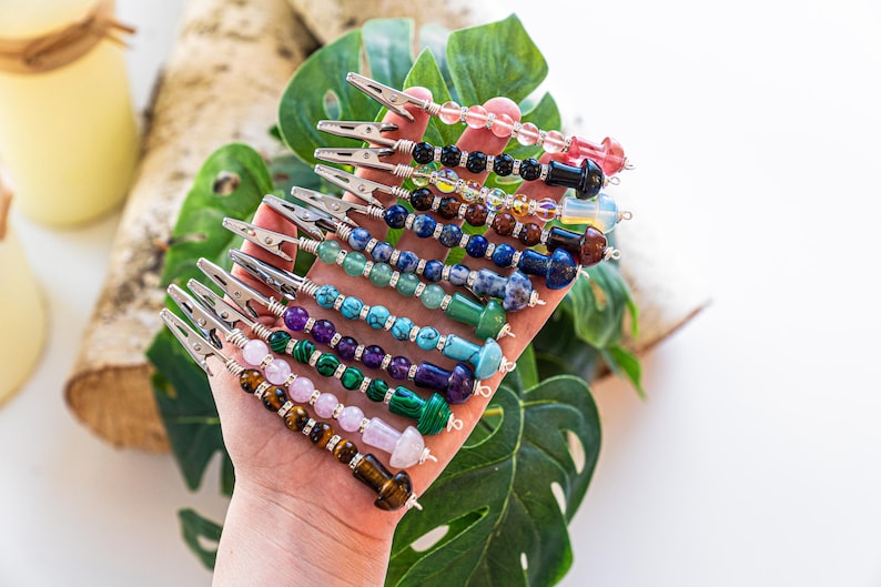 May include: A hand holding a set of 10 colorful beaded clips with silver metal clips. Each clip has a different combination of beads, including pink, blue, green, brown, and black beads.