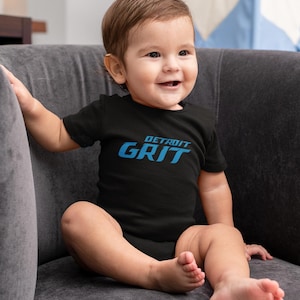 May include: A baby wearing a black bodysuit with the text "DETROIT GRIT" in blue lettering. The baby is sitting on a gray couch.